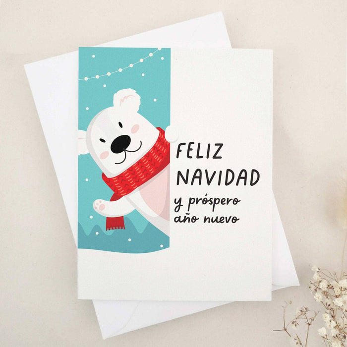 Christmas card with “Feliz Navidad y Próspero Año Nuevo” in bold black text, illustrated with a smiling polar bear in a red scarf against a snowy blue background with festive string lights, paired with white envelope — XOXOKristen.