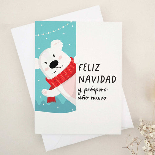 Christmas card with “Feliz Navidad y Próspero Año Nuevo” in bold black text, illustrated with a smiling polar bear in a red scarf against a snowy blue background with festive string lights, paired with white envelope — XOXOKristen.