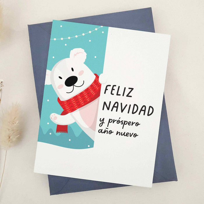 Holiday card featuring polar bear in cozy red scarf on winter blue background with festive lights, paired with Spanish greeting, styled with blue envelope — XOXOKristen.