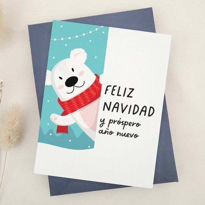 Holiday card featuring polar bear in cozy red scarf on winter blue background with festive lights, paired with Spanish greeting, styled with blue envelope — XOXOKristen.