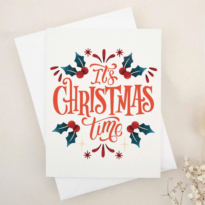 Christmas card on slightly textured off-white cardstock with bold red-orange “It’s Christmas Time” lettering, surrounded by dark green holly leaves and red berries, paired with white envelope — XOXOKristen.
