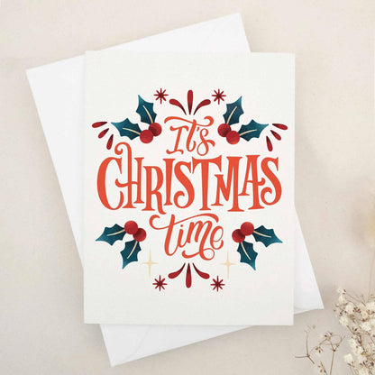 Christmas card on slightly textured off-white cardstock with bold red-orange “It’s Christmas Time” lettering, surrounded by dark green holly leaves and red berries, paired with white envelope — XOXOKristen.