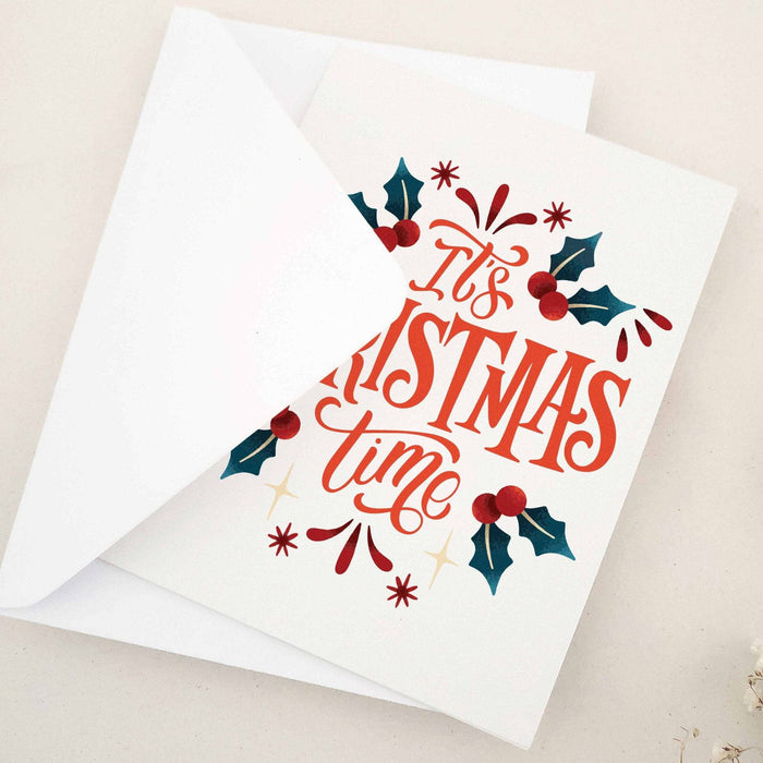 Folded holiday card with “It’s Christmas Time” in decorative script, framed by festive holly and berry accents, displayed with matching envelope — XOXOKristen.