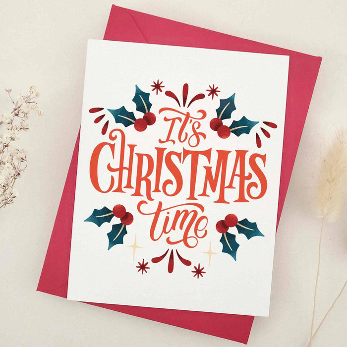 Standing Christmas card on off-white cardstock with red-orange calligraphy and holly leaf border, styled with red envelope — XOXOKristen.