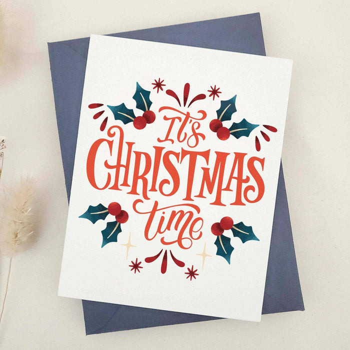 Holiday card featuring “It’s Christmas Time” in bold lettering, framed by greenery and berry details, displayed with coordinating blue envelope — XOXOKristen.