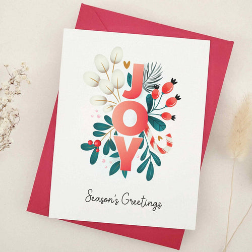 Christmas card on slightly textured off-white cardstock with bold “JOY” letters decorated in floral motifs, red berries, pine sprigs, and greenery, paired with envelope — XOXOKristen.