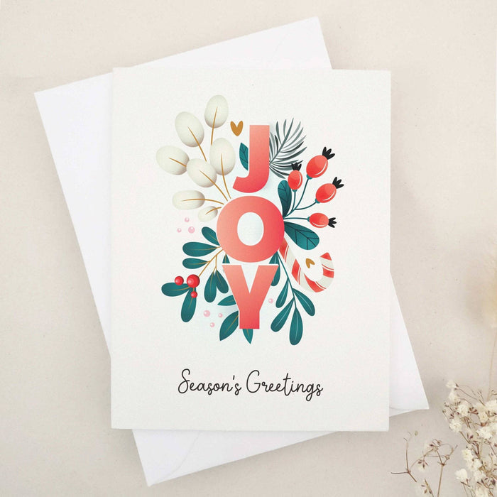 Folded holiday card featuring the word “JOY” in coral red letters surrounded by soft cream leaves, berries, and foliage, with “Season’s Greetings” below — XOXOKristen.