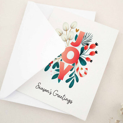 Christmas card partially open, front showing festive “JOY” floral lettering with candy-cane stripe details, greenery, and red berries — XOXOKristen.