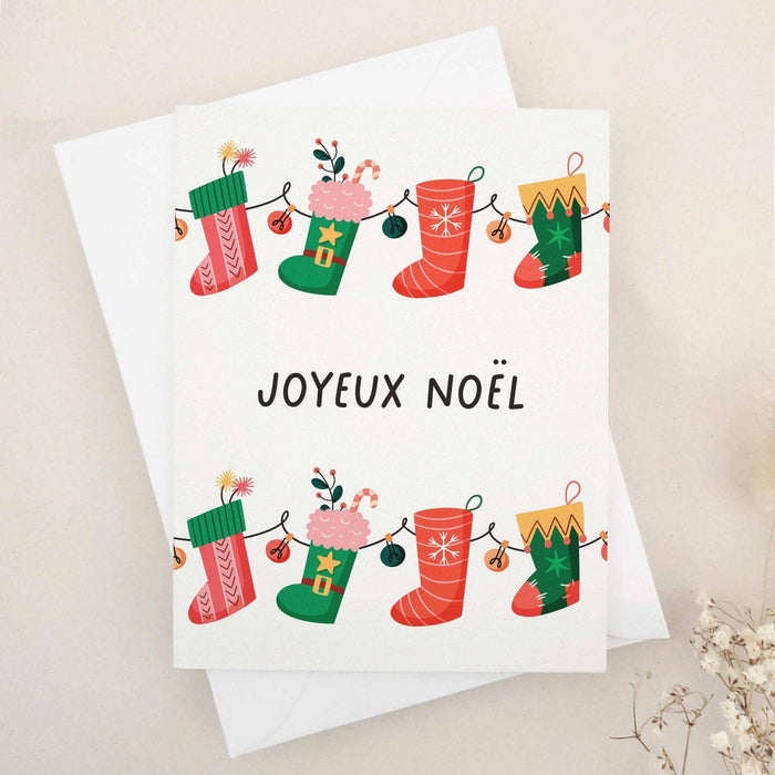 Christmas card with “Joyeux Noël” in bold black text, decorated with a garland of red and green stockings filled with gifts, candy canes, and florals, paired with white envelope — XOXOKristen.