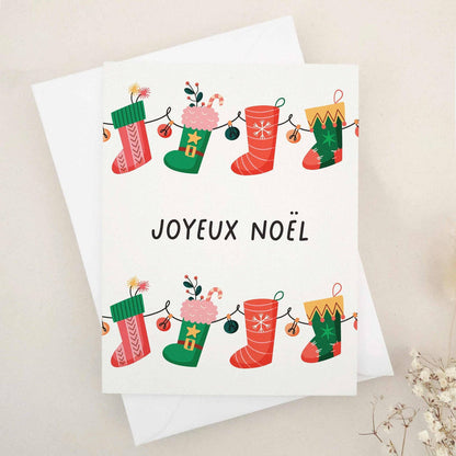 Christmas card with “Joyeux Noël” in bold black text, decorated with a garland of red and green stockings filled with gifts, candy canes, and florals, paired with white envelope — XOXOKristen.