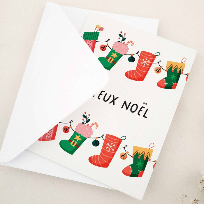 Folded holiday card featuring “Joyeux Noël” below a line of festive stockings filled with presents and greenery, displayed with envelope — XOXOKristen.