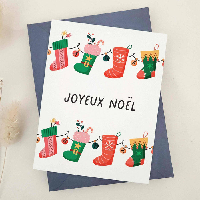 Holiday card featuring colorful hanging stockings, gift boxes, and candy cane accents above “Joyeux Noël” text, styled with blue envelope — XOXOKristen.