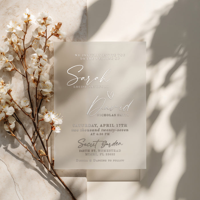 A semi-transparent vellum wedding invitation printed in metallic silver foil, placed on a softly textured stone background. The warm natural lighting highlights the reflective details of the foil and the frosted translucency of the vellum. The composition evokes luxury and minimalism, ideal for sophisticated weddings with a neutral color palette. – XOXOKristen
