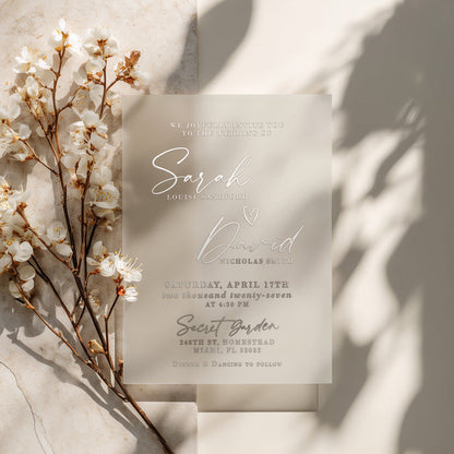 A semi-transparent vellum wedding invitation printed in metallic silver foil, placed on a softly textured stone background. The warm natural lighting highlights the reflective details of the foil and the frosted translucency of the vellum. The composition evokes luxury and minimalism, ideal for sophisticated weddings with a neutral color palette. – XOXOKristen