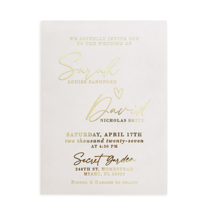 A vertical display of a translucent vellum wedding invitation featuring luminous gold foil lettering, positioned upright among natural stone and dried floral stems. The soft sunlight and gentle shadows enhance the reflective golden accents and the frosted vellum texture, creating a refined, romantic mood. The composition captures the modern luxury of personalized wedding stationery. – XOXOKristen
