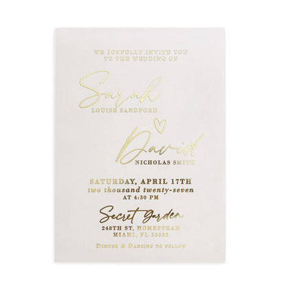 A vertical display of a translucent vellum wedding invitation featuring luminous gold foil lettering, positioned upright among natural stone and dried floral stems. The soft sunlight and gentle shadows enhance the reflective golden accents and the frosted vellum texture, creating a refined, romantic mood. The composition captures the modern luxury of personalized wedding stationery. – XOXOKristen