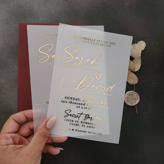 A vertical display of a translucent vellum wedding invitation featuring luminous gold foil lettering, positioned upright among natural stone and dried floral stems. The soft sunlight and gentle shadows enhance the reflective golden accents and the frosted vellum texture, creating a refined, romantic mood. The composition captures the modern luxury of personalized wedding stationery. – XOXOKristen