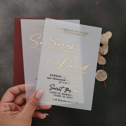 A vertical display of a translucent vellum wedding invitation featuring luminous gold foil lettering, positioned upright among natural stone and dried floral stems. The soft sunlight and gentle shadows enhance the reflective golden accents and the frosted vellum texture, creating a refined, romantic mood. The composition captures the modern luxury of personalized wedding stationery. – XOXOKristen